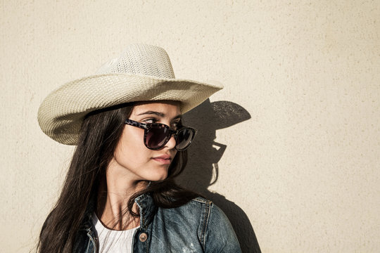 portrait of girl in sunglasses in cowboy hat on background of wall, serious look to side
