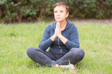 Mature beautiful active happy woman in the morning in the park, relax after sports exercises.  Middle woman in the yoga pose sits on the grass outdoor.