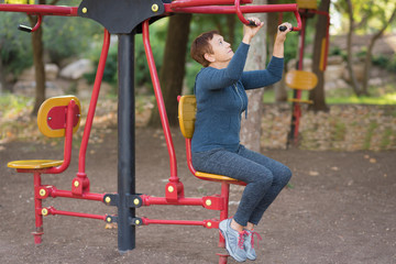 Fototapeta na wymiar Senior woman working out on the sports public equipment in the outdoor gym.A sportive active elderly woman doing physical exercise in a park at sunny day. 