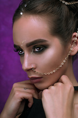 Girl with cool fashionable make-up posing in studio,matte lips, smokey gold eyes, piercing chain from nose over purple dark background with magic glow. Holiday disco celebration