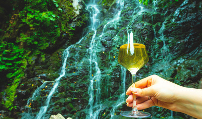 female hand holding a glass of white wine in the background rock mountain with a waterfall