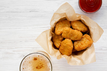 Chicken nuggets in a paper box, ketchup and glass of cold beer on a white wooden surface, top view. Overhead, from above, flat lay. Copy space.
