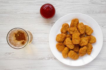 Chicken nuggets on a white plate, ketchup and glass of cold beer on a white wooden surface. Overhead, from above, flat lay.
