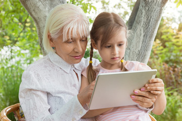 Grandmother with her granddaughter studying a book on a tablet, work on the tablet. Granny at the age of 70 years old with great granddaughter in her arms. Concept. Relationship in the family. 