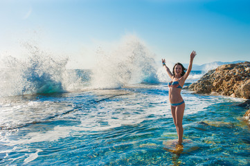 A girl in a swimsuit, splashing from the waves, Brunette girl rejoices splashing from the waves. Young happy woman jumping with raised arms, waving a blue bikini swimsuit in a blue swimsuit and a