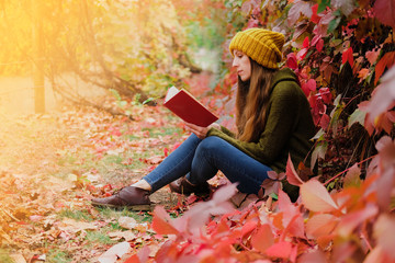 Girl in mustard yellow knit hat and marsh green wool sweater sitting among colorful ivy in autumn and reading a paper book