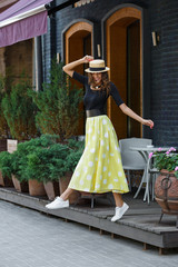 Fototapeta premium Stylish brunette girl dressed in black blouse and long yellow in white peas skirt walks in the city street on a sunny day