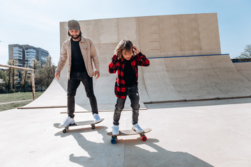 Father and his little son dressed in the casual clothes ride skateboards in a skate park with...