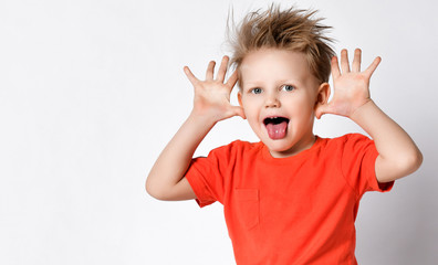 Grimacing kid boy in orange t-shirt shows big ears with his hands and put his tongue at us