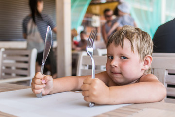 A little boy holds a knife and fork and waits for food. A hungry child is sitting at a table in a...