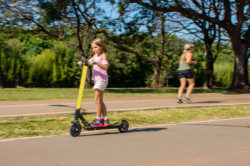 Child playing with scooter in city park and adults walking.