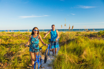 Summer people lifestyle happy couple biking on beach relaxing outdoors activity at sunset. Young woman and man riding recreational bikes bicycles on USA Florida holidays getaway.