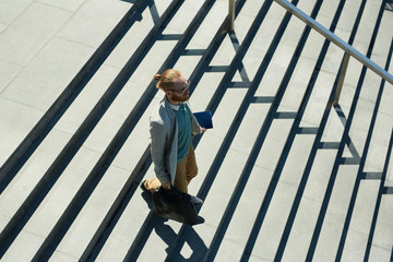Above view of serious confident hipster employee with red beard carrying briefcase and diary while...