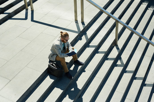Concentrated creative bearded manager in sunglasses sitting on step of stairs and writing ideas down in diary