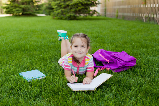 child girl schoolgirl elementary school student  lying on the grass and draws in a notebook. concept back to school. outdoor activities