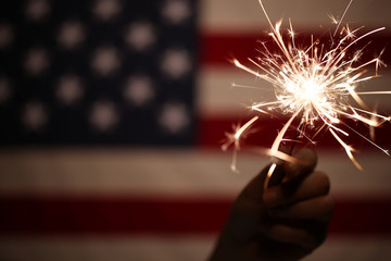 Hand holding lit sparkler in front of the American Flag for 4th of July celebration