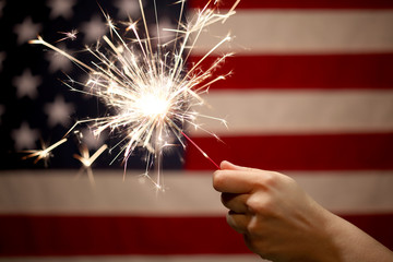 Hand holding lit sparkler in front of the American Flag for 4th of July celebration