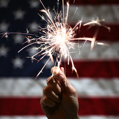 Hand holding lit sparkler in front of the American Flag for 4th of July celebration