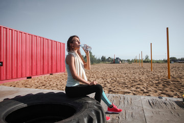 Close up portrait of young pretty caucasian fitness woman sitting on the tractors tire outdoor and drinking water. Breaking relax while exercise workout. Concept of health and sport lifestyle. 