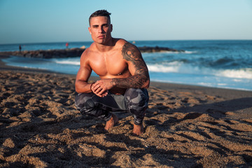 Tattooed bodybuilder sexy male coach at the beach. He sits and poses for the camera and shows his muscles