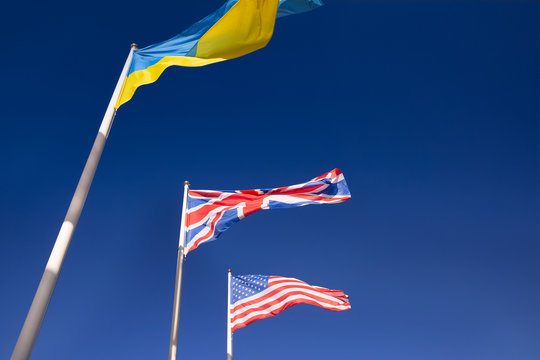 Flags Of Ukraine, Britain And United States Of America Are Developing In Wind. Three Flags On Flagpoles Against Blue Sky. Concept Of Support And Cooperation Of Ukraine With Britain And United States.