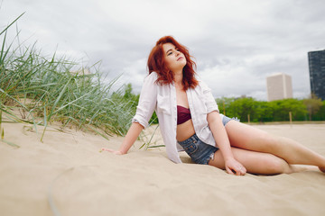 A young redheaded girl in a white shirt and jeans shorts sitting on the sand on the beach