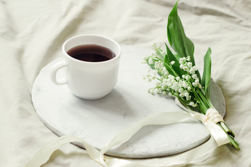 delicate bouquet of fresh lilies of the valley decorated with silk ribbon and freshly brewed coffee on a wooden tray on the bed