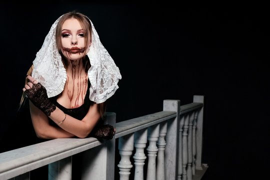 Concept halloween makeup. A sexy girl dressed as a nun stands on a balcony near a white marble railing. Black background. Copy space.