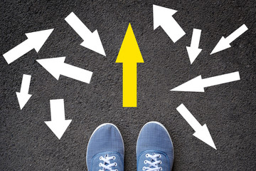 Fototapeta premium Confusing direction arrows on asphalt ground, feet and shoes on floor, personal orientation concept