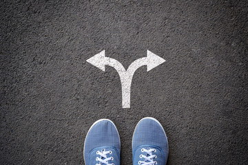 Pair of feet standing on tarmac road with arrow print pointing in two different directions for the concept of making decision at crossroad.