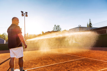 Preparing a tennis court for the match. A man is watering a field with a hose.