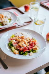 Plate of salad with tomatoes, jamon and baked vegetables on the white table. Glass of iced water with mint and lemon.