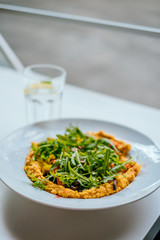 Bowl of carrot vegan hummus with chickpea, caramelized carrot, mashrooms, garlic, paprika, tahena, cumin, rocket salad and baked cauliflower. Glass of iced water with mint and lemon.