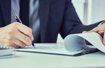 Hand of businessman in suit filling and signing with blue pen partnership agreement form clipped to pad closeup. Management training course, some important document, team leader ambition concept.