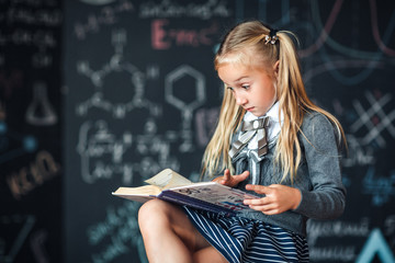 Little girl blonde in school uniform looks at the book with a surprised face. Chalkboard with school formulas. Complex school program, does not match the age of the child.