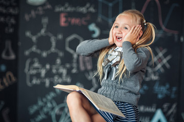 A sweet blond girl schoolgirl sits on a chalkboard with school formulas background. Works homework by reading a book Amazed to put his hands on his head, opening his mouth wide.