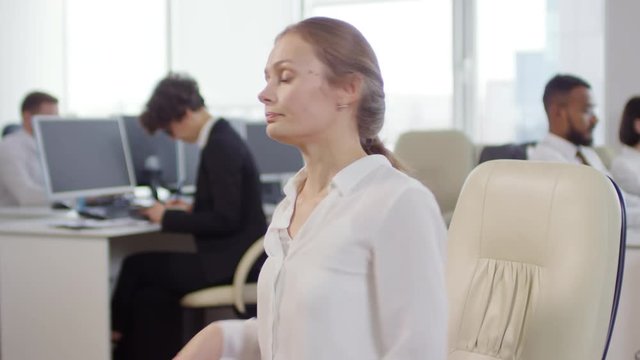 Medium shot of pretty Caucasian businesswoman sitting on office chair, stretching her back, shoulders and neck while her coworkers working on computers on background
