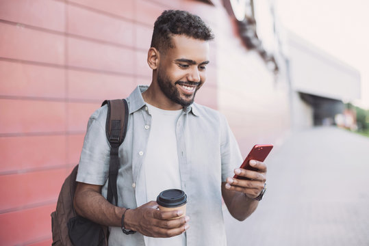 Young handsome man using smartphone in a city. Cheerful men holding mobile phone and coffee. Travel, lifestyle, technology concept