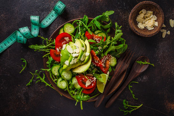 Green salad with avocado, tomatoes, cucumbers and nuts in wooden plate, centimeter on dark background. Fitness and diet food concept.