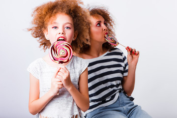 Mom and daughter with natural small curls on the head with bright lollipops, on a light background. Space for text.