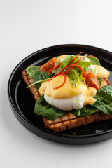 Side close view of grilled italian sandwich on wheat toast with greens, benedict egg, chili pepper and cherry tomatoes, served on black dishware and isolated on white