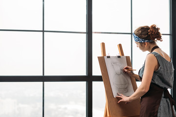 Art class. Side view of young lady drawing sketch of vase in studio. French window background. Copy...