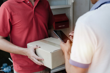 young man customer appending signature in digital mobile phone receiving parcel post box from courier with home delivery service man smiling face in red uniform at home, express delivery concept