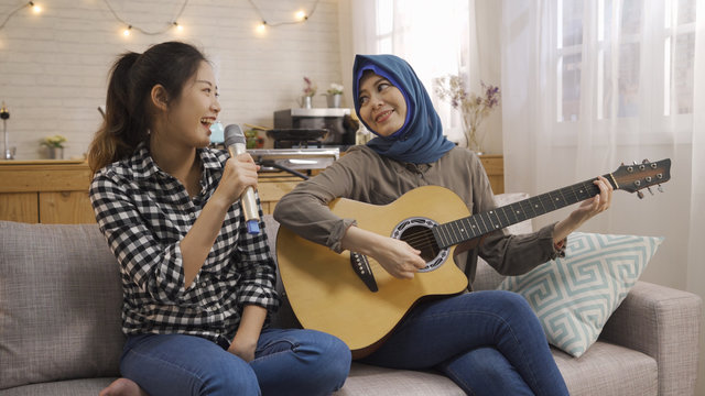 Cheerful happy group of multi ethnic friends having fun in dormitory practice songs for performance. two young asian student college girls chinese and muslim playing guitar singing sitting on couch.