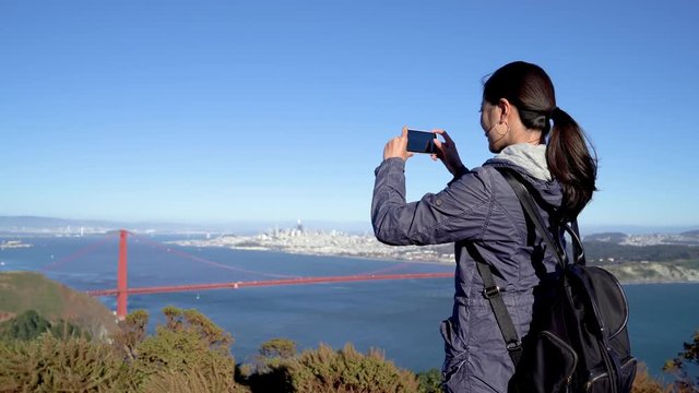 Young chinese woman hiker stand on top of mountains take photo of landscape on cellphone in holiday summer vacation travel trip in san francisco usa sightseeing red golden gate bridge. memory record 