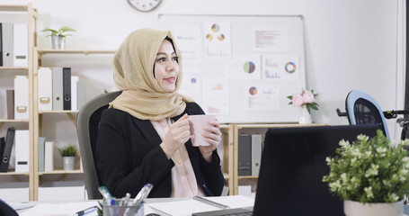 Attractive young arabic businesswoman sitting at workplace desk in office relax having break. malay lady drinking coffee holding tea cup and smiling looking aside with joy emotion. copy space.