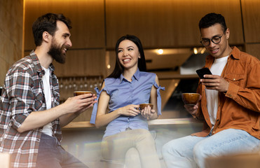 Smiling friends drinking coffee in cafe. Multiracial hipsters communication together, laughing, discussing creative ideas, holding cups of tea and mobile phone