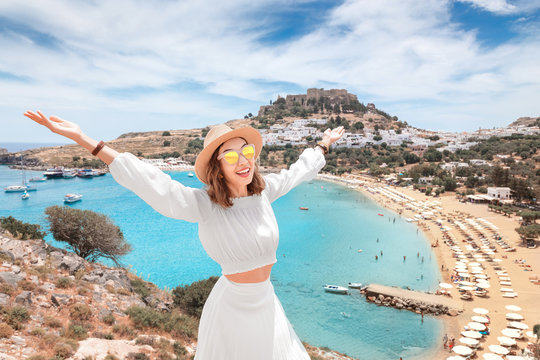 Happy asian woman raised hands travels in Greece. Having fun at the stunning view on sea resort and old town of Lindos on the mountain