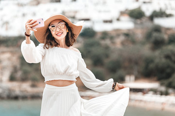 Elegant tourist girl taking selfie photo on her smartphone at the background of ancient white...