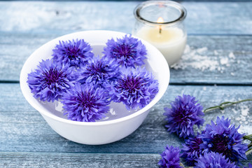 Fototapeta na wymiar blue cornflowers lie in water in a white bowl near a burning candle against the background of blue old boards.